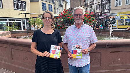 Ein Mann und eine Frau halten einen Handzettel in der Hand. Sie stehen auf dem Marktplatz in Butzbach, im Hintergrund ein Brunnen.